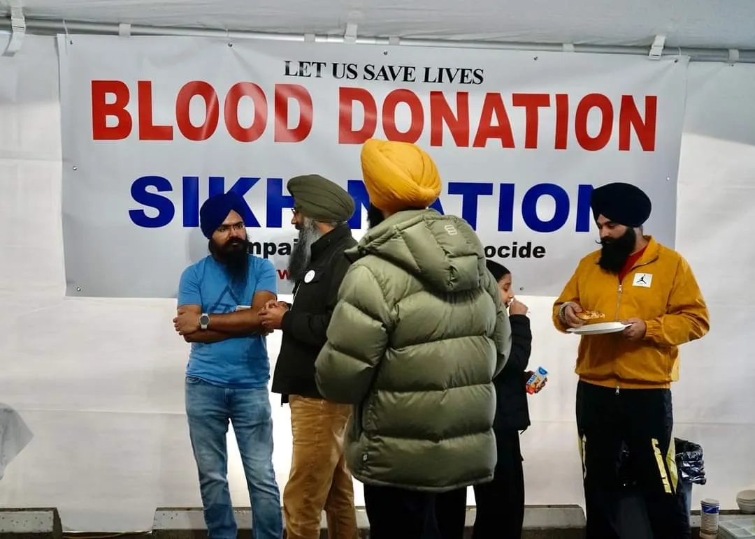 Volunteers standing and talking in front of a large banner reading 'Let us save lives — Blood Donation — Sikh Nation' at one of the camps.