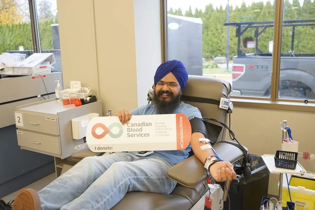 Daljot Singh smiling in a donation chair at a Canadian Blood Services clinic, holding an 'I donate' paddle, with a pressure cuff and line on his outstretched arm.