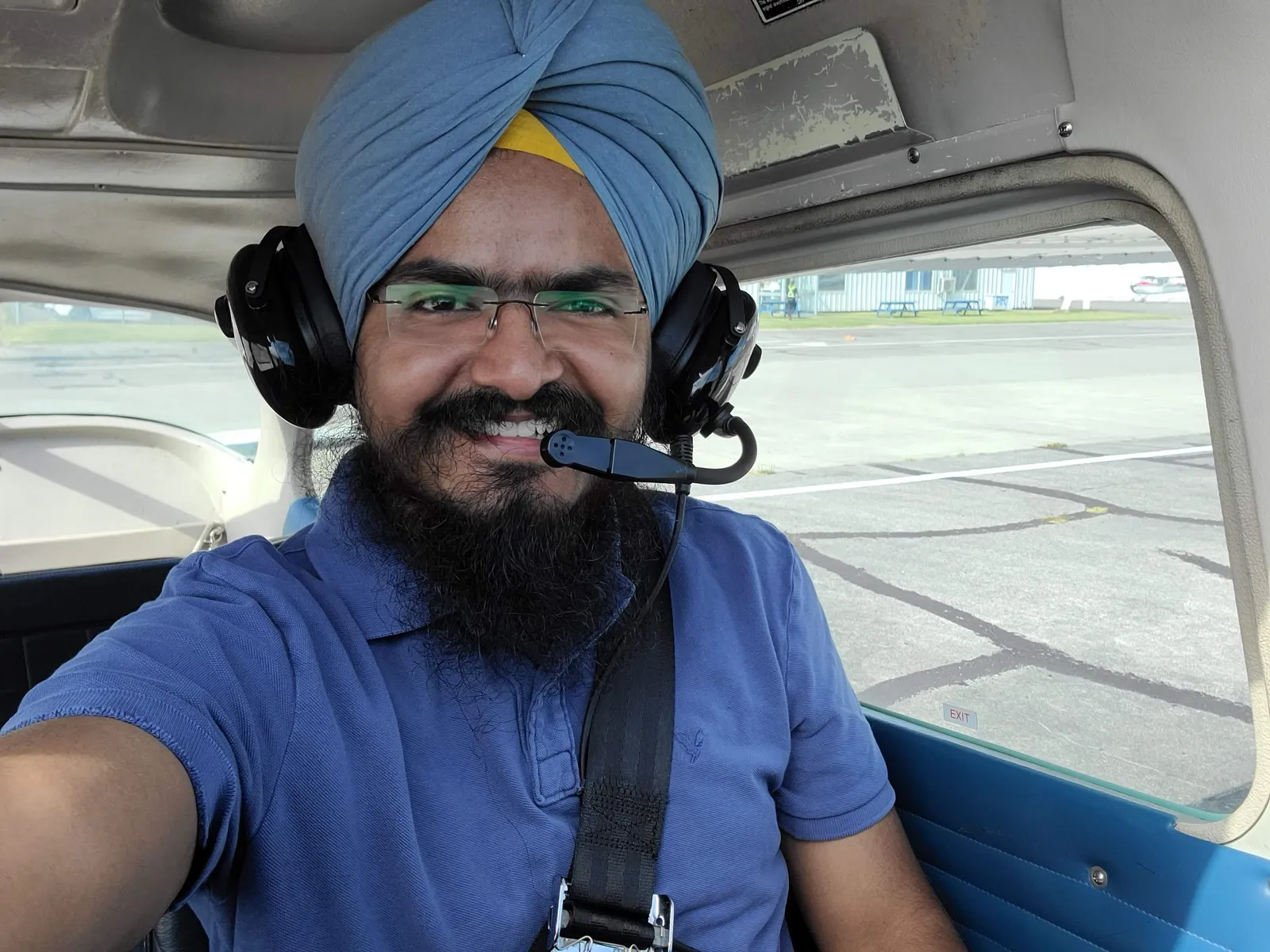 Daljot Singh grinning in the left seat of a small aircraft, aviation headset on, the airfield visible through the window.