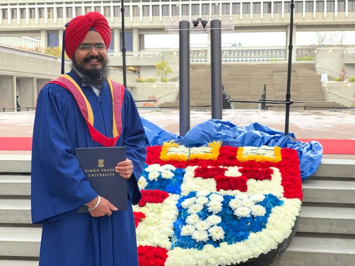 Daljot Singh in blue and red convocation robes holding a Simon Fraser University diploma folder on the Burnaby campus.