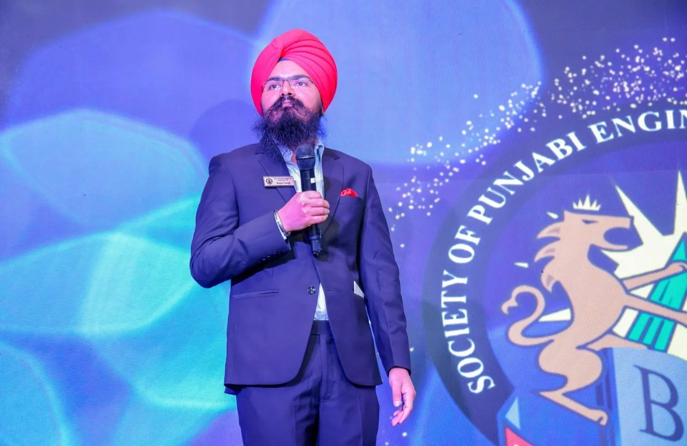 Daljot Singh in a navy suit and red turban speaking into a microphone on stage, the Society of Punjabi Engineers and Technologists of BC crest projected behind him.