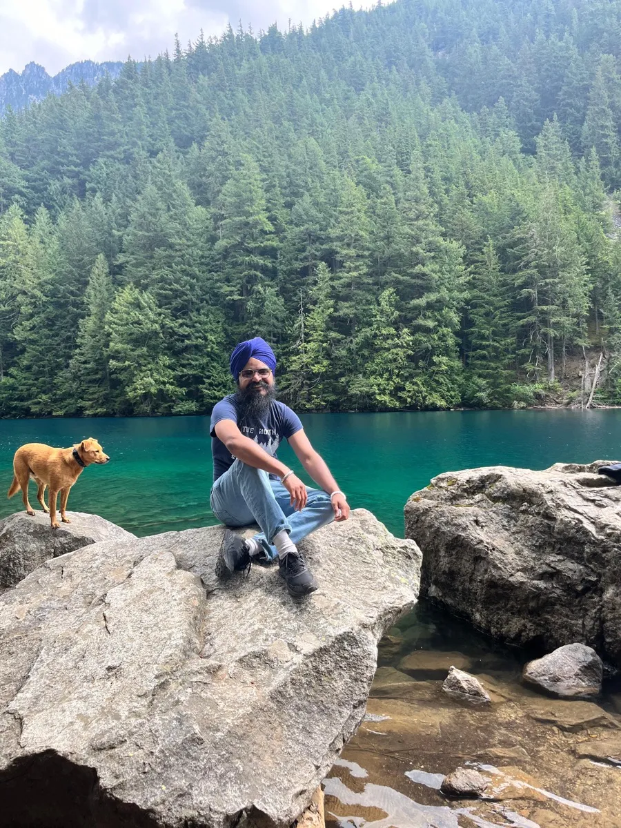 Daljot Singh sitting on a boulder beside a turquoise alpine lake ringed with fir trees, a golden dog standing on the rock beside him.