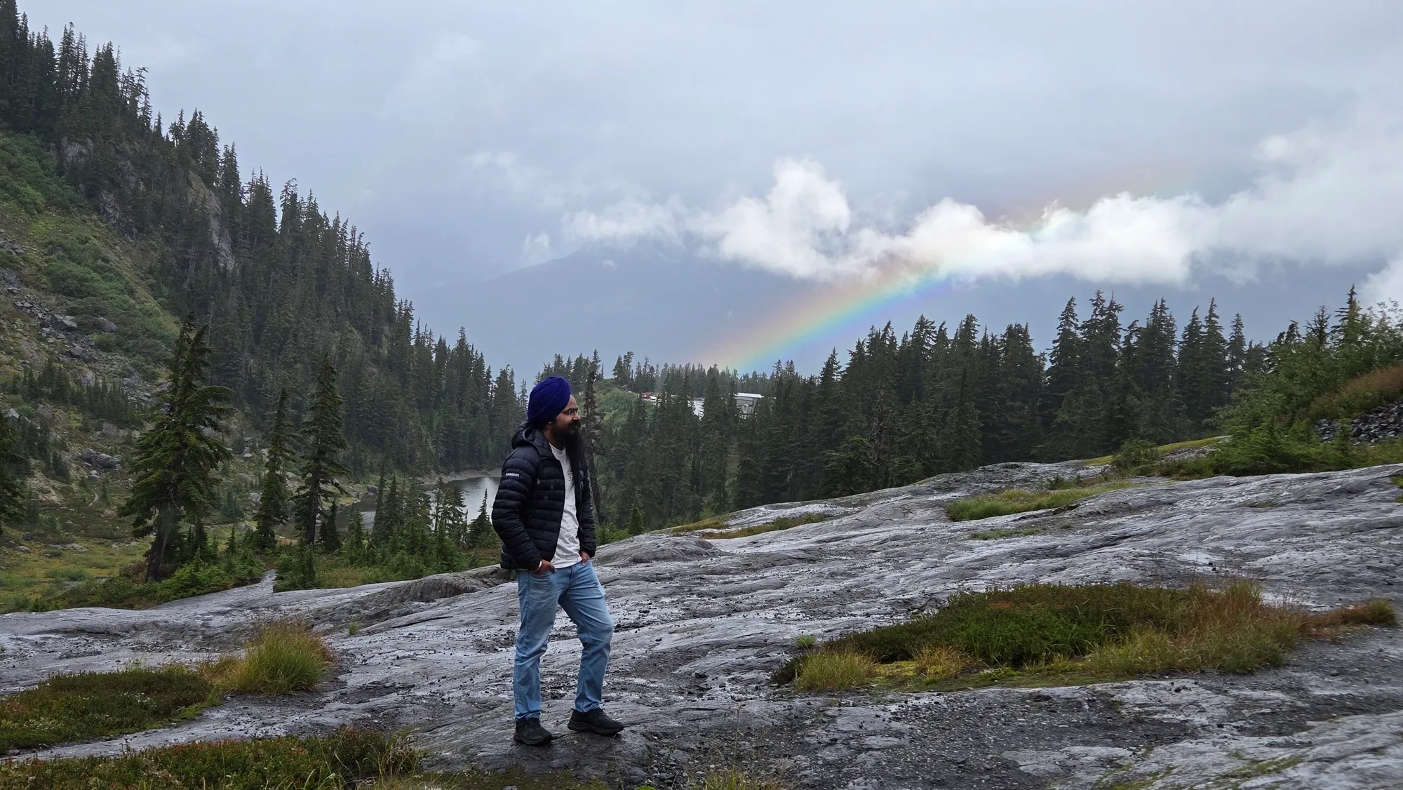 Daljot Singh walking across wet granite slabs below a mountain ridge, a rainbow breaking over the treeline behind him.