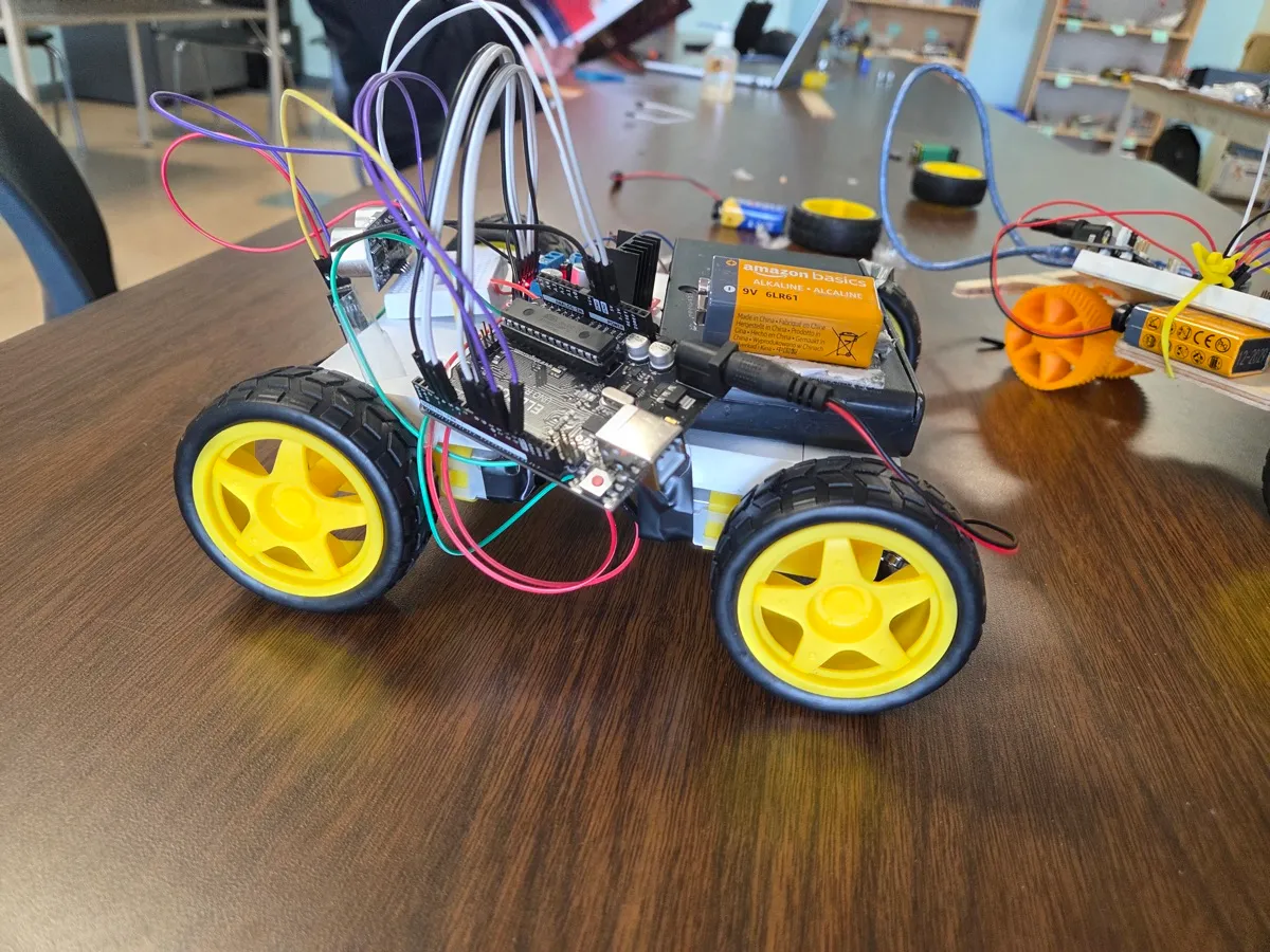 A student-built four-wheeled robot on a classroom table: an Arduino board, a battery pack and a tangle of jumper wires above bright yellow wheels.