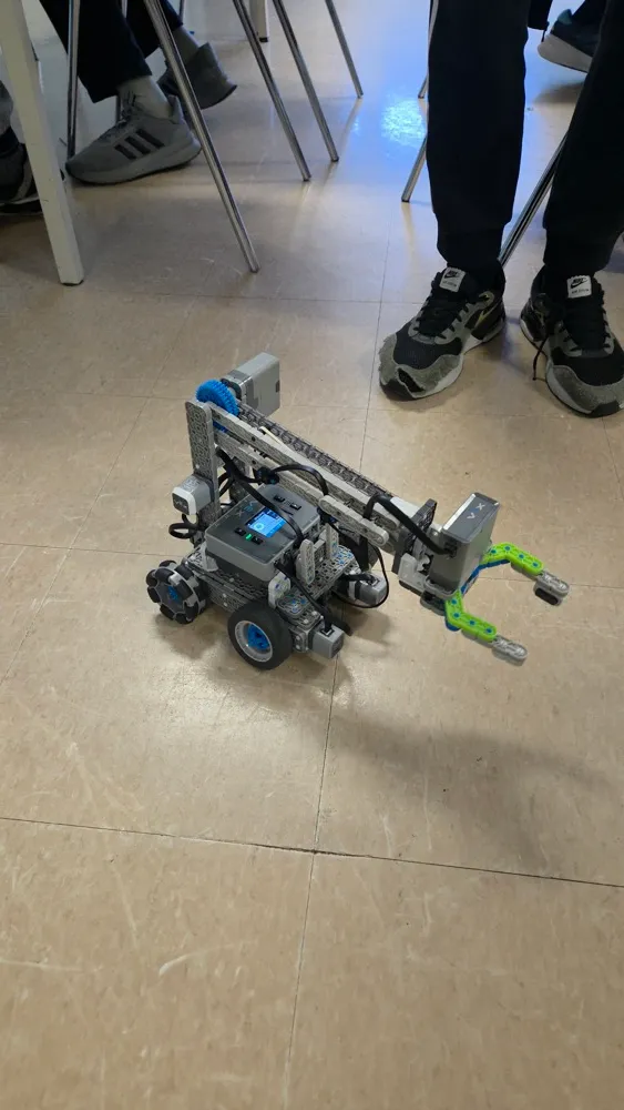 A LEGO robot with a green gripper claw on a classroom floor, students' shoes visible in the background.