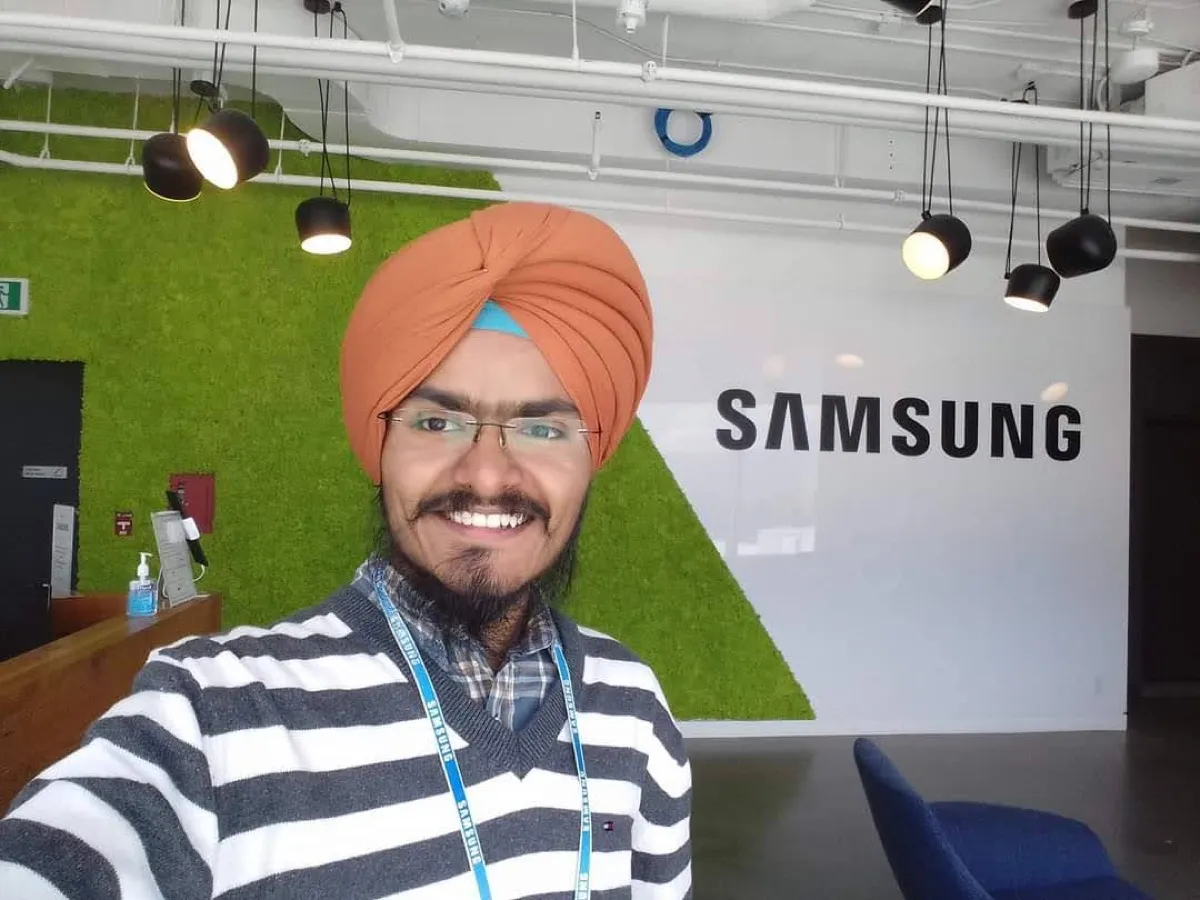 A younger Daljot Singh in an orange turban with a company lanyard, standing in front of a large Samsung sign in an office.