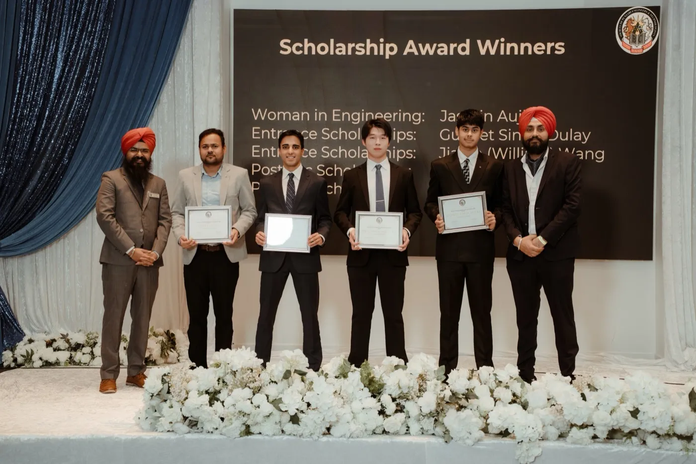 Four scholarship recipients holding certificates on stage beneath a screen reading Scholarship Award Winners, with Daljot Singh standing at the far left.