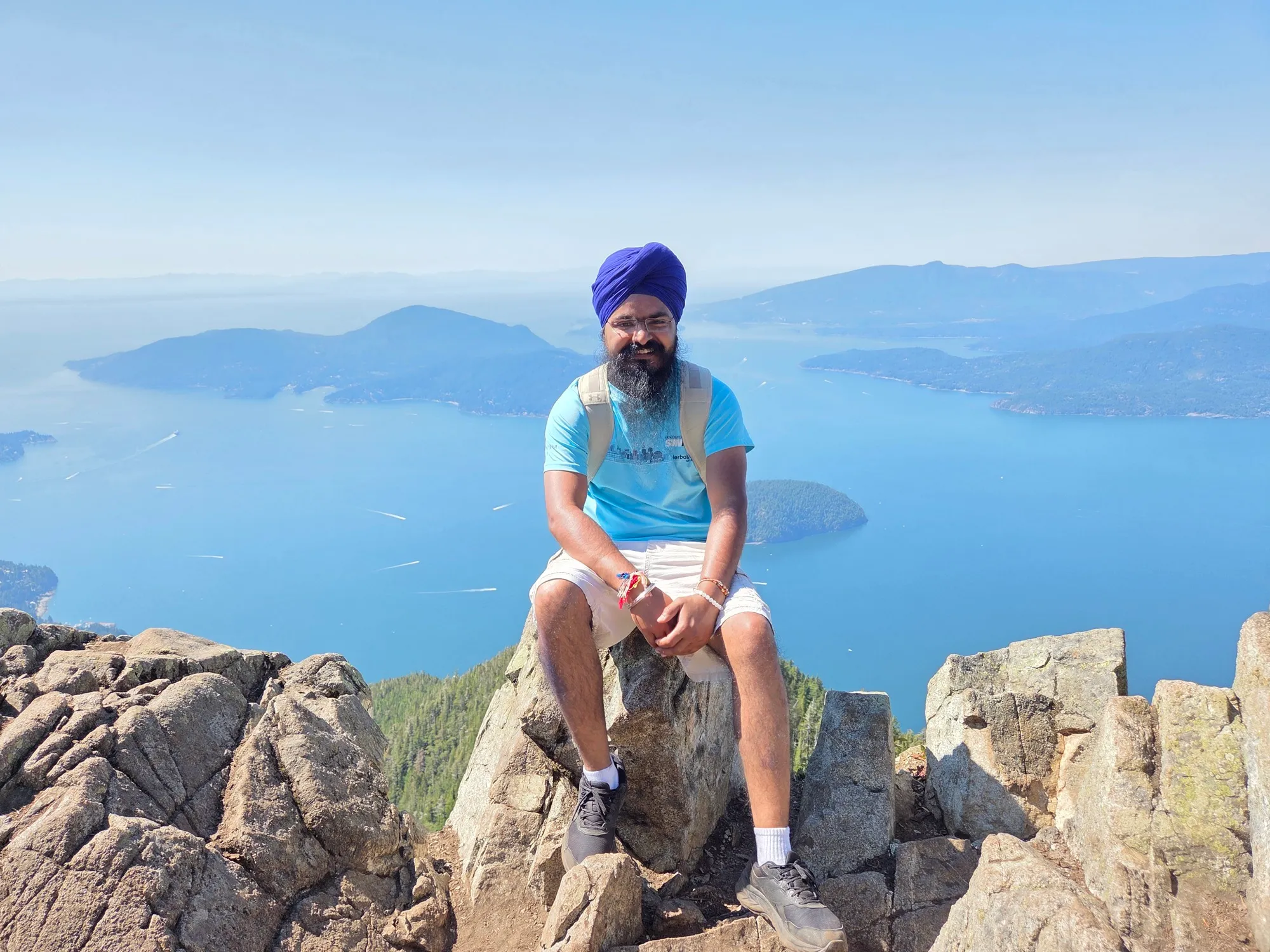 Daljot Singh sitting on a rocky summit high above Howe Sound, islands and blue water stretching out behind him.